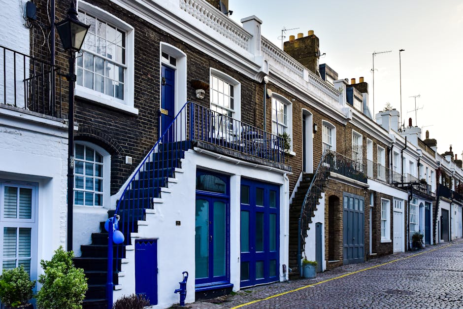 Exterior view of a row of traditional residential houses in Fulham, London, featuring brick facades with white window frames and contrasting blue doors and stair railings. The houses are built in a terraced style, with small balconies and exterior stairs leading to upper floors. The pavement in front is cobbled, and some greenery is visible in planters and small bushes near the entrances. The scene is illuminated by natural daylight, highlighting the clean and well-maintained appearance of the properties, reflecting the importance of surface cleaning and maintenance promoted by Cleaners Fulham in their domestic cleaning services.