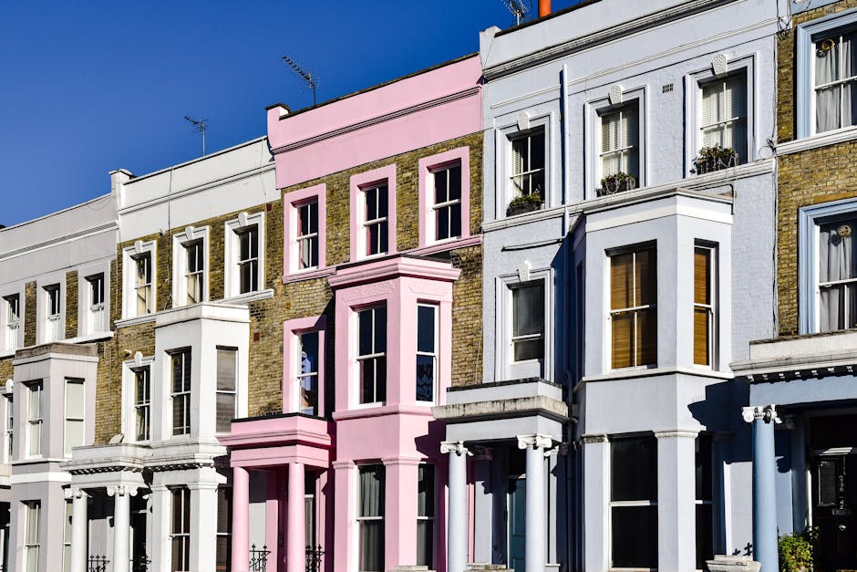 A row of Victorian-style residential buildings with mixed brick and painted facades, featuring large sash windows and decorative cornices. The buildings have varying pastel and neutral color schemes, including white, light pink, and blue. Some windows are adorned with flower boxes and curtains visible inside. The facades appear clean and well-maintained against a bright blue sky, with sunlight casting soft shadows. This image exemplifies building exteriors that could benefit from comprehensive cleaning and maintenance, as offered by Cleaners Fulham, specialists in domestic cleaning and surface sanitisation.