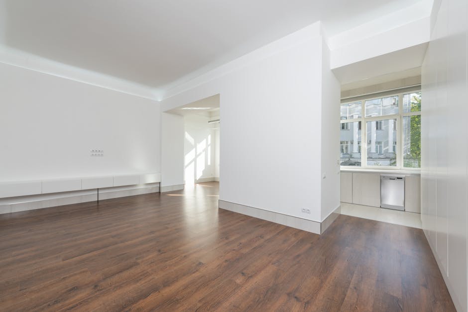 A spacious, empty living room with white painted walls and a wooden floor that appears thoroughly cleaned and polished, reflecting natural light from large, multi-pane windows on the right side. The room features a built-in white storage bench along the left wall and a corner area with a small alcove housing window blinds and a radiator below the windows. The surfaces, including the glossy white walls and clean wooden floor, are free of dust or debris, indicating a comprehensive surface cleaning and sanitisation process. The room's bright and airy atmosphere is enhanced by modern white trim and ceiling molding, with visible electrical outlets on the walls. This image exemplifies professional domestic cleaning aimed at maintaining hygiene and a pristine living environment, as offered by Cleaners Fulham, specializing in deep cleaning and surface maintenance for properties like North End Road flats, Fulham.