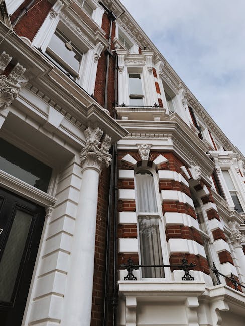 Photograph of a row of elegant white Victorian-style townhouses on North End Road in Fulham, featuring ornate architectural details, large windows, and small balconies with black wrought iron railings. The street is illuminated by traditional street lamps, and a leafless tree with bare branches stands prominently in front of the buildings. Several parked cars are visible along the curb, and the scene is bathed in natural daylight with a clear blue sky overhead, showcasing a clean and well-maintained residential area. This image highlights the classic London architecture consistent with the SW6 postcode, reflecting the environment where professional cleaning services by Cleaners Fulham are often required for maintaining property hygiene and presentation.